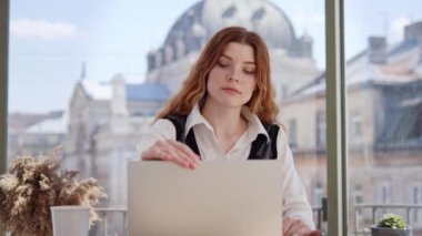 Güzel, çekici, kahverengi saçlı, beyaz tişörtlü iş kadını laptopunu açıyor ve panoramik pencereli bir ofiste kameraya gülümsüyor antik şehir manzarası sunuyor.