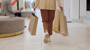 Alışveriş meraklısı bir kadının arkası, elinde alışveriş torbalarıyla alışveriş merkezinde geziniyor. Memnun müşteri, güzel alışveriş merkezinin koridorunda dizüstü bilgisayar kullanan bir adamın yanından geçiyor.