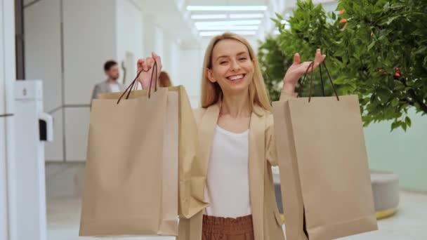 在购物中心购物后 穿着白色T恤和夹克的漂亮 微笑的女孩拿着装有衣服的手袋 在后台 有一对年轻夫妇站着 — 图库视频影像
