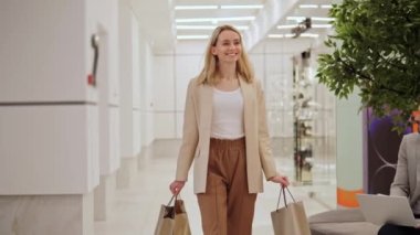 Mutlu, güzel, neşeli sarışın kız alışveriş çantalarını elinde tutuyor ve alışveriş merkezinde dolaşıyor. Kadın gülümser, etrafında döner ve insanlar arka plandayken alışverişlerinden zevk alır.