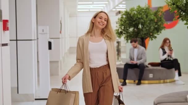 美しく 楽しいブロンドの女の子は手にショッピングバッグを保持し ショッピングセンター モールの周りを歩きます 女性は笑顔 彼女の購入を楽しみ 余暇を過ごし 店で服を買う — ストック動画