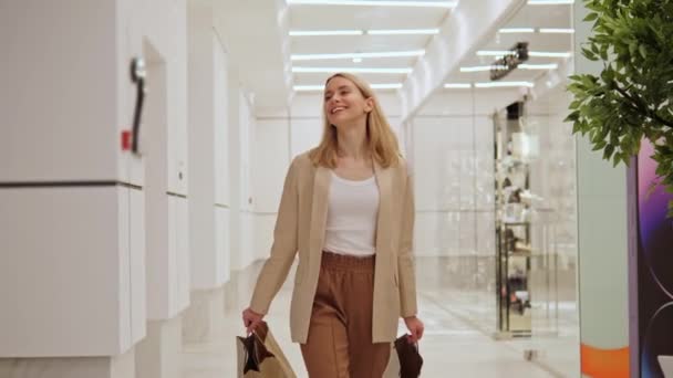明るく 美しく 楽しいブロンドの女の子は手にショッピングバッグを持っており ショッピングセンターの周りを歩いています 女性の笑顔 見て回る 彼女の購入の人々を楽しんでいる背景にある販売の季節 — ストック動画