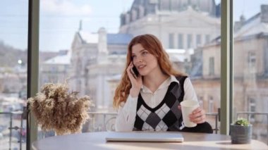 Pozitif kızıl saçlı iş kadını birden fazla işte çalışıyor cep telefonu üzerinden dizüstü bilgisayarla konuşuyor Cafe City 'deki müşteriyle çevrimiçi proje çözüyor ya da iş arkadaşlarıyla konuşuyor.