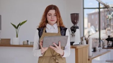 Kızıl saçlı bayan satıcı, önlük giymiş küçük bir kafe sahibi ve beyaz tişörtlü, elinde tablet olan bir kahve dükkanı siparişi alıyor. Kahvenin içi parlak, rahat ve ferah.