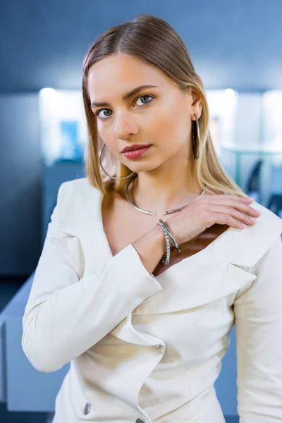 Enchanting and beautiful blonde model showcases various diamond accessories on her body in a jewelry boutique. The girl is wearing gold earrings, a diamond ring, and a sparkling gold necklace