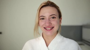 Karizmatik ve doğal güzelliğe sahip güzel bir kız, diş teli ve beyaz bir bornoz giyiyor, güzellik prosedürlerinden sonra bir güzellik kliniğinde karbon soyma ve cilt gençleştirme gibi kameraya gülümsüyor.
