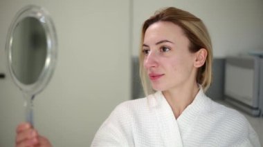 Doğal güzelliği olan güzel bir kız, elinde bir ayna tutuyor, beyaz bir bornoz giyiyor, güzellik prosedürlerinden sonra kendine bakıyor, mesela karbon soyma ve cilt yenileme gibi, güzellik kliniğinde.