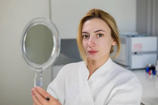 Naturally beautiful positive girl, without makeup, dressed in a white robe, smiles at the camera following beauty treatments like carbon peeling and skin rejuvenation, at a beauty clinic