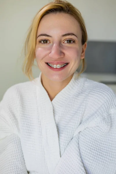 Positive girl with natural beauty face with out make up, wearing braces and a white robe, smiles at the camera after beauty procedures, skin rejuvenation, in a beauty clinic, spa salon