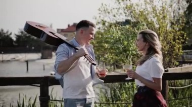 Mavi tişörtlü, elinde gitar olan bir adam göl kenarında bayan arkadaşıyla konuşuyor.