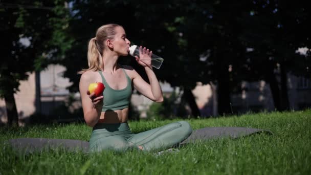 健康的女孩坐在一个荷花的姿势里 从一个运动瓶子里喝水 手里拿着苹果 — 图库视频影像