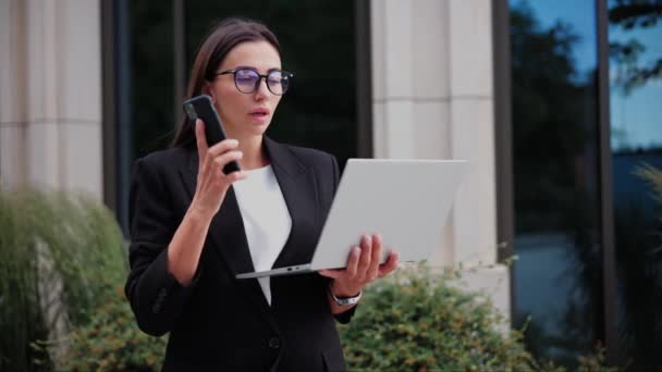 オフィスビルの近くの携帯電話で話している眼鏡で忙しいかわいいビジネスマン ファッションフォーマルスーツを着ているビジネス女性は 現代のラップトップコンピュータを保持しながら携帯電話で話しています — ストック動画