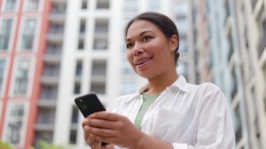 Olumlu gülümseyen bayan dışarıda elinde akıllı telefonuyla dikiliyor. Çekici Afrikalı Amerikalı Kadın Cep Telefonu Kullanarak Sohbet Ediyor. Açık havada Çekici Kadın Mesajlaşması