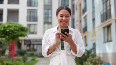 Gülümseyen Mutlu Kız Açık havada Smartphone kullanıyor. Genç Afrikalı Amerikalı Kadın Kaydırma Videoları veya Sosyal Medya Fotoğrafları. Çevrimiçi Çalışan Gülümseyen Kadın Alışveriş İçin Modern Akıllı Telefon Kullanıyor