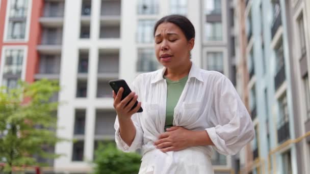 アフリカ系アメリカ人女性は スマートフォンを使用しながら不快感を感じ 病気やクランプを感じます 救急車を呼ぶ外側に立っているスタマキャッシュの若いサディレディ 健康コンセプトの問題 — ストック動画