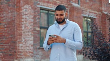 Genç yaşta kaygısız Hintli bir adam şehir sokağının ortasında duruyor ve online mobil uygulama kullanarak kız arkadaşıyla sohbet ediyor. Sıradan kıyafetli sakallı bir adam elinde akıllı bir telefon tutuyor ve başını çeviriyor..