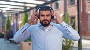 Koyu saçlı, sakallı, kablosuz kulaklık takan ve kameraya gülümseyen yakışıklı Hintli adam. Gökyüzü mavisi gömlekli karizmatik genç yetişkin favori müziğin tadını çıkarıyor..