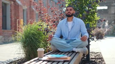 Hintli erkek meditasyon ustası, elinde kahve ve kitaplarla tahta bankta otururken nilüfer çiçeği pozu egzersizi yapıyor. Kotlu ve mavi gömlekli barışçıl bir adam açık havada gözleriyle dengede duruyor..