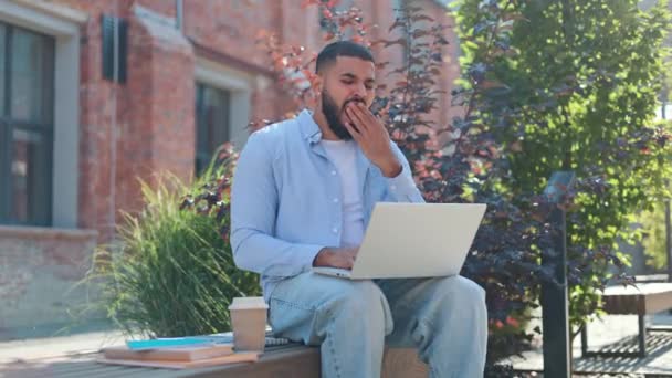 英俊的阿拉伯男子坐在木制长椅上 手里拿着咖啡和书本 在现代无线笔记本电脑上工作 男青年粗心大意地穿衣 感到精疲力尽 打呵欠和关闭便携装置 — 图库视频影像