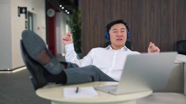 Розслаблений Азіатський Чоловік Сидить Ногами Офісному Столі Слухаючи Улюблену Пісню — стокове відео