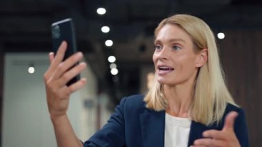 Çekici beyaz iş kadınının portresi profesyonel çalışma alanında modern akıllı telefondan video görüşmesi yapıyor. Takım elbiseli şık bir kadın enerjik bir şekilde konuşuyor ve el kol hareketleri yapıyor..