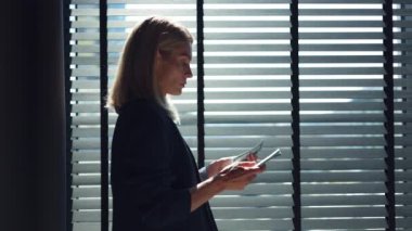 Pencerenin yanında duran, yatay jaluzili ve dolar banknotları sayan kadın ofis çalışanının yan görüntüsü. Beyaz tenli resmi giysili sarışın aylık maaşının toplam miktarını kontrol ediyor..