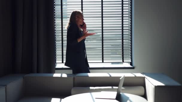 站在窗口时 穿着时髦西装的愤怒女性在与同事的移动对话中大喊大叫 在情感上做手势 阳光透过敞开的百叶窗照射在黑暗的办公室 — 图库视频影像