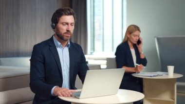 Kablosuz kulaklıkla ofis masasında oturan yakışıklı sakallı adam ve video konferansı için dizüstü bilgisayar kullanıyor. Kafkasyalı iş kadını. Sarı saçlı. Arka planda cep telefonundan konuşan..