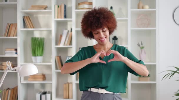 魅力的なブルネット女性は ハートジェスチャーを手で示しながら幸せでロマンチックです アフリカ系アメリカ人女性 優しい気持ちを表現し 明るいオフィスで元気よく踊る — ストック動画
