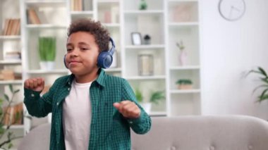 Etnik kökenli aktif bir çocuk. Kablosuz kulaklıkla müzik dinliyor ve ferah bir odada canlı dans ediyor. Enerjik çocuk evde eğleniyor. Çocuklar ve modern teknoloji kavramı.