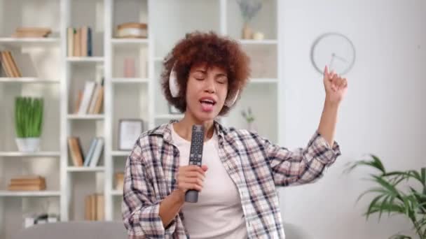 アフリカ系アメリカ人のエネルギッシュな女性は リモートコントローラーで屋内でリラックスして歌います リズム的に体を動かし ドライブミュージックを楽しむイヤホンを身に着けているコンテンツの女性 — ストック動画