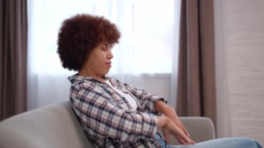 Yorgun, Afro-Amerikalı bir kadının rahat koltukta el ele tutuşup gözlerini kapamasının yan görüntüsü. Kıvırcık saçlı genç bir kadın hafta sonları evde dinleniyor..