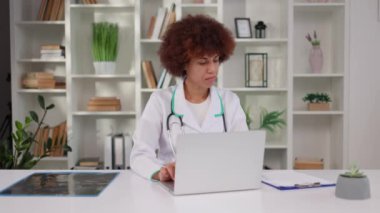 Laboratuvar önlüklü Afrikalı Amerikalı kadın terapist modern klinikte otururken hasta bilgilerini internete yüklemek için kablosuz laptop kullanıyor. Tıbbi sağlık ve teknoloji konsepti.