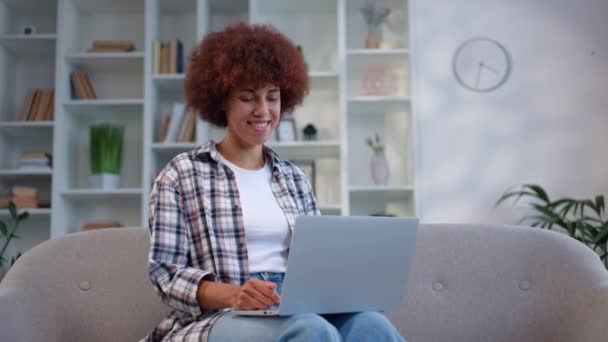 年轻的非洲裔美国女性坐在舒适的沙发上 参加关于现代小玩意的在线会议 有魅力的女人穿着休闲装 用笔记本电脑和同事聊天 — 图库视频影像