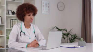Çok ırklı pratisyen hekimin hastasıyla hastanede iş yerinde otururken bilgisayarında video görüşmesi var. Tıbbi üniformalı genç bir kadın online ziyarete gidiyor, gülümsüyor ve el sallıyor..
