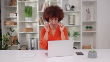 Portakal rengi bluzlu Afro-Amerikalı kadın, modern iş yerinde masa başında kablosuz bilgisayarla otururken gözlük takıyor. Kıvırcık saçlı, gülümseyen ve kameraya bakan çekici bir kadın..