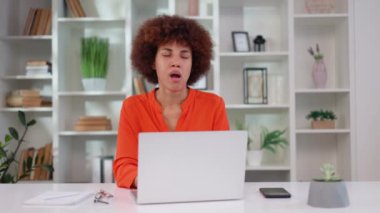 Yorgun bir Afrikalı Amerikalı kadın modern dizüstü bilgisayarla ofis masasında oturuyor ve esniyor. Şık turuncu bluz giyen, bitkin ve bitkin düşmüş bir kadın şirket çalışanı..