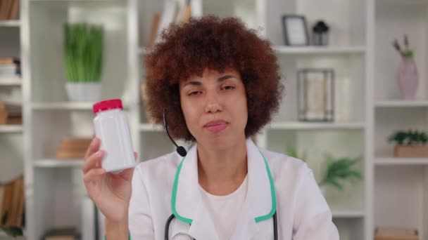 非洲裔美国女人戴着无线耳机 一边在医疗柜与病人在线交谈 一边展示着一罐白色药丸 电视预约期间提供药物建议的女治疗师 — 图库视频影像