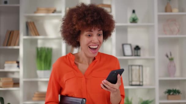スマートフォンの画面を見ながら幸せを表現する多民族女性社員 オレンジブラウスの感情的なダークハイレッド女性は 職場での誠実な感情で良いニュースに反応します — ストック動画