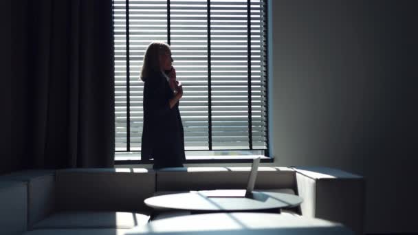 一个穿着西装的严肃的高加索女人站在办公室窗前 用现代智能手机聊天的侧视图 在与伴侣的紧张对话中 自信的女士在感情上做手势 — 图库视频影像