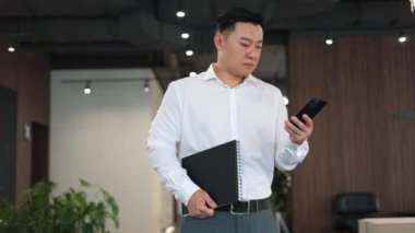 Odaklanmış satış müdürü elinde siyah defter tutuyor ve müşteriden gelen akıllı telefondan online başvuru üzerinden alınan belgeleri inceliyor. Takım elbiseli Asyalı profesyonel atölyede modern cihaz kullanıyor..
