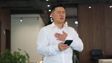 Stresli ofis çalışanı çalışma yerinde dikilirken ve akıllı telefon kullanırken göğüs ağrısı çekiyor. Asyalı bir uzman nefes darlığı, mide bulantısı ve mide ekşimesi hissediyor. pf sağlık sorunları kavramı.