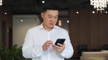 Borsaya bakarken akıllı telefon uygulamasında yükselen grafikleri izleyen mutlu Asyalı tüccarın ön görüntüsü. Resmi giyinmiş şok olmuş bir adam heyecan verici duygular ifade ediyor ve başarıyı kutluyor..