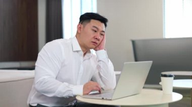Çağdaş çalışma alanında yuvarlak masaüstünde otururken Asyalı uyruklu bir ofis çalışanı sıkılıyor. Resmi olarak giyinmiş, yorgun erkek işçi avuç içine yaslanmış ve taşınabilir bilgisayarda daktilo kullanıyor..