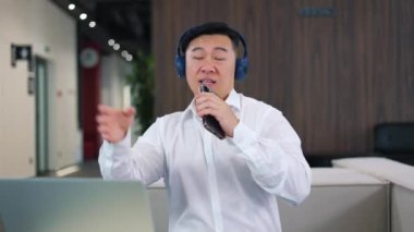 Asya uyruklu neşeli erkek CEO 'su kablosuz kulaklıkla müzik dinliyor ve modern akıllı telefonuyla karaoke yapıyor. Geniş bir ofisteki bilgisayar işinden alıkoyan yakışıklı adam..