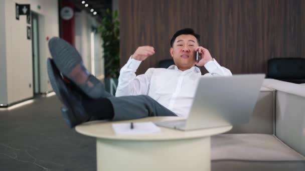 Sonriente Hombre Asiático Hablando Teléfono Inteligente Moderno Mientras Descansa Sofá — Vídeos de Stock