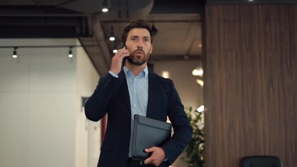 黒いフォルダを握り 企業のオフィスビルを歩いている深刻な顔の表情を持つ自信のあるビジネスマン クライアントとスマートフォンで会話する成功した起業家 — ストック動画