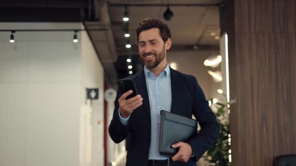 オフィス廊下を歩き回り 携帯電話の画面を見る成功した雇用主のフロントビュー 笑顔の男は ソーシャルメディアでスクロールラインをビジネス服を着て 次に目をそらす — ストック動画