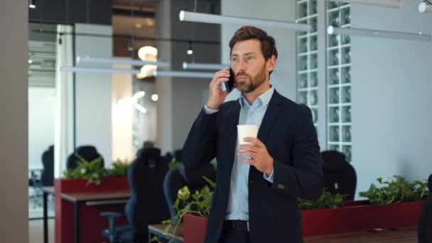 現代のコーポレートオフィスに立っている間 コーヒーを飲み スマートフォンで話すハンサムなひげ付き男 パートナーとの会話を持つスタイリッシュなフォーマルスーツを着たコーカサス実業家 — ストック動画
