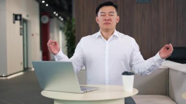 Olgun Asyalı adam, ofis masasında meditasyon ve mudra hareketleriyle rahatlıyor. Beyaz yakalı iş adamı, iş günü kablosuz bilgisayarda enerji ve uyum kazanıyor..